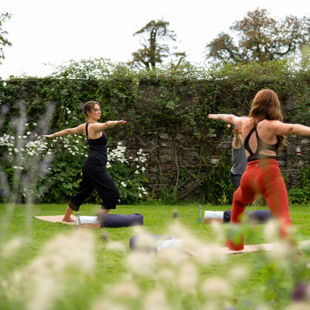 Drie mensen beoefenen yoga buiten op gras, staand op matten in de Krijger II-houding. Twee vrouwen staan tegenover elkaar met gestrekte armen, terwijl een man ertussen zijwaarts staat. Weelderig groen, bloemen en een stenen muur op de achtergrond creëren een vredige omgeving. | Echt Ierland Vakanties