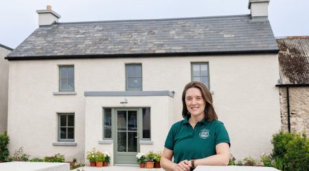 Een vrouw met lang bruin haar, gekleed in een groene polo en spijkerbroek, glimlacht terwijl ze voor een gerenoveerde boerderij staat. Het huis heeft crèmekleurige muren, een grijs leien dak en bloempotten langs het pad dat naar de voordeur leidt. | Echt Ierland Vakanties