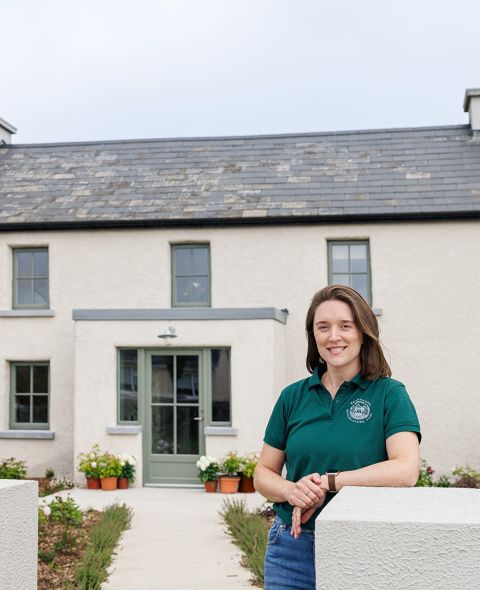 Een vrouw met lang bruin haar, gekleed in een groene polo en spijkerbroek, glimlacht terwijl ze voor een gerenoveerde boerderij staat. Het huis heeft crèmekleurige muren, een grijs leien dak en bloempotten langs het pad dat naar de voordeur leidt. | Echt Ierland Vakanties
