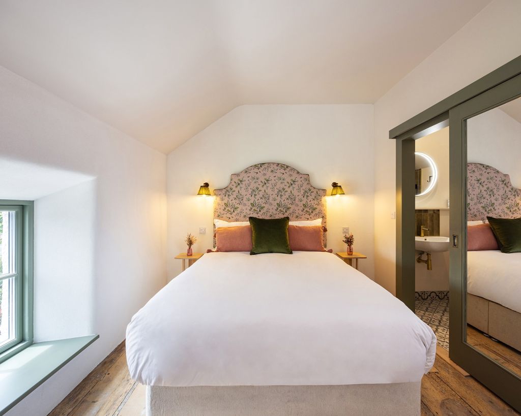 Een knusse slaapkamer met een groot bed met een met bloemen bekleed hoofdbord, wit linnen en roze en groene accentkussens. Twee kleine ronde bijzettafeltjes met bloemen, wandlampen aan beide kanten en een spiegel met groene omlijsting weerspiegelt de wastafel. Natuurlijk licht komt binnen via een raam. | Echt Ierland Vakanties