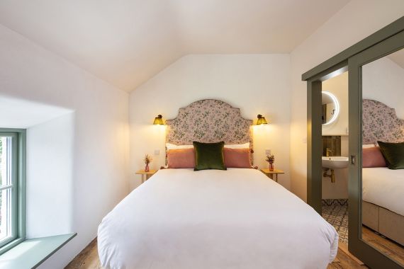 Een knusse slaapkamer met een groot bed met een met bloemen bekleed hoofdbord, wit linnen en roze en groene accentkussens. Twee kleine ronde bijzettafeltjes met bloemen, wandlampen aan beide kanten en een spiegel met groene omlijsting weerspiegelt de wastafel. Natuurlijk licht komt binnen via een raam. | Echt Ierland Vakanties