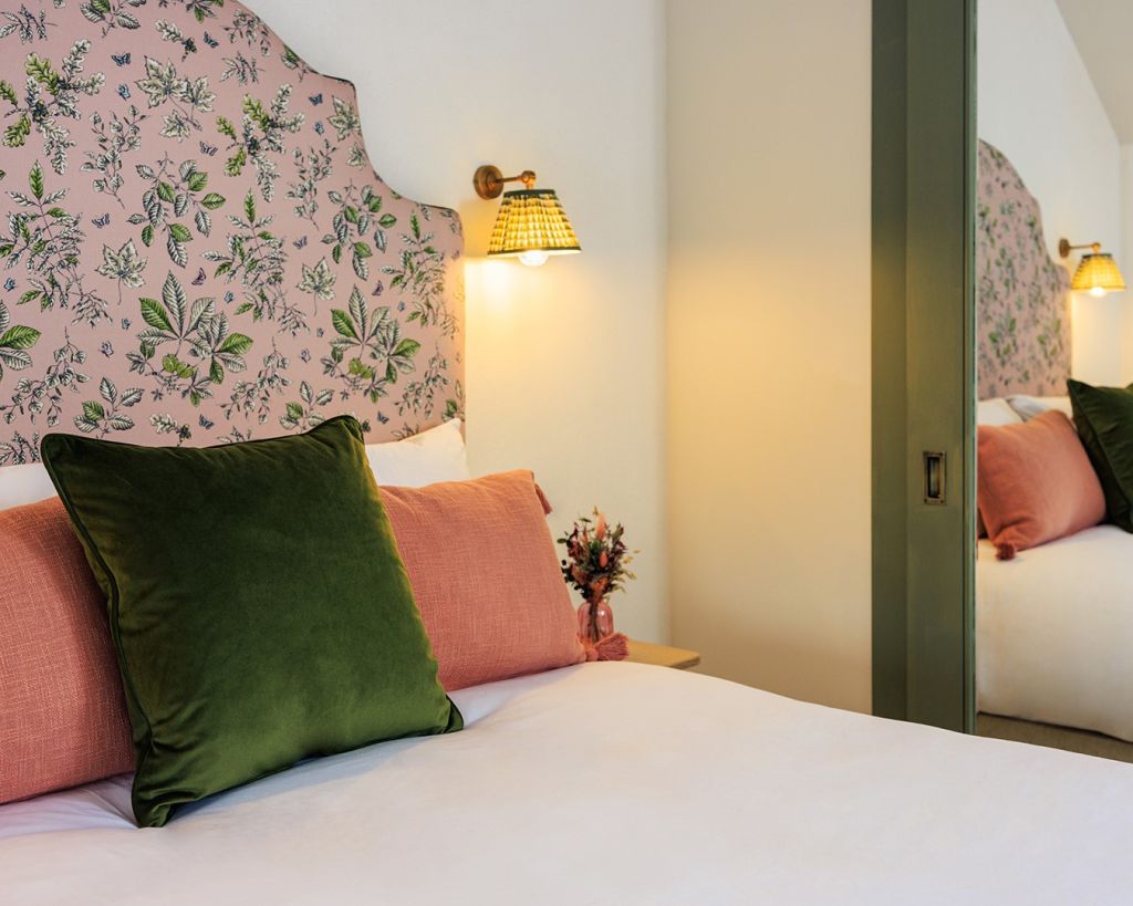 Een knus bed met een roze, met bloemen bekleed hoofdbord, een groen fluwelen kussen en twee roze kussens. Op het nachtkastje staat een klein boeketje. Een messing wandlamp met een geweven lampenkap straalt warm licht uit en een grote spiegel reflecteert een deel van het bed en de kussens. | Echt Ierland Vakanties