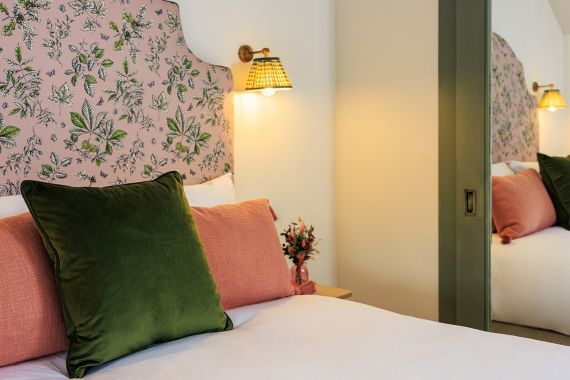 Een knus bed met een roze, met bloemen bekleed hoofdbord, een groen fluwelen kussen en twee roze kussens. Op het nachtkastje staat een klein boeketje. Een messing wandlamp met een geweven lampenkap straalt warm licht uit en een grote spiegel reflecteert een deel van het bed en de kussens. | Echt Ierland Vakanties