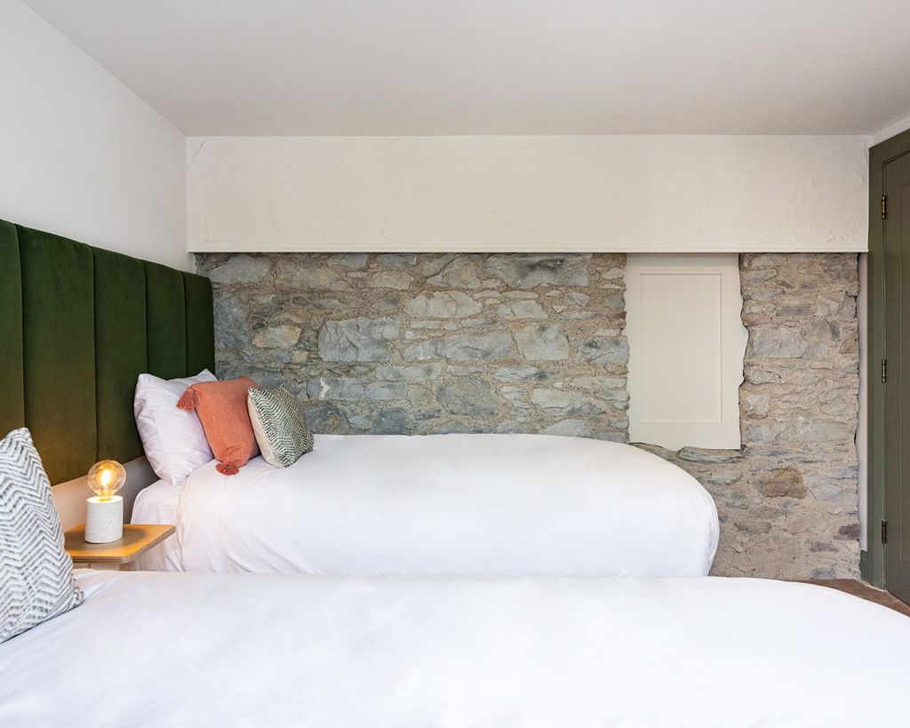 Een moderne slaapkamer met twee witte bedden, groene gestoffeerde hoofdborden, een stenen accentmuur en een klein houten nachtkastje met een gloeilamp en een kussen met zigzagpatroon. De kamer heeft een minimalistische, gezellige sfeer met accenten in aardetinten. | Echt Ierland Vakanties