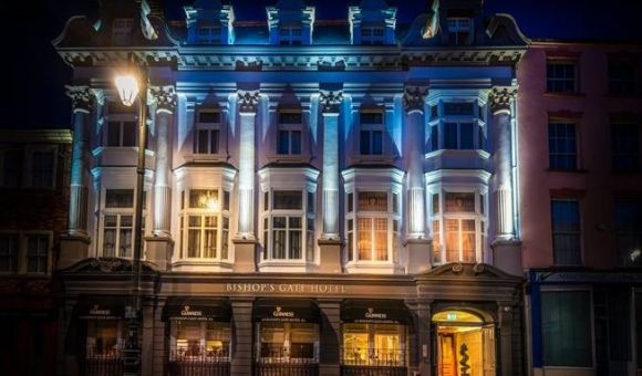 De sierlijke gevel van het Bishop's Gate Hotel, 's avonds verlicht, is voorzien van hoge ramen, decoratieve zuilen en zachtblauwe verlichting. Op de begane grond hangen Guinness-borden en warm verlichte entrees, die contrasteren met de donkere straat buiten. | Echt Ierland Vakanties