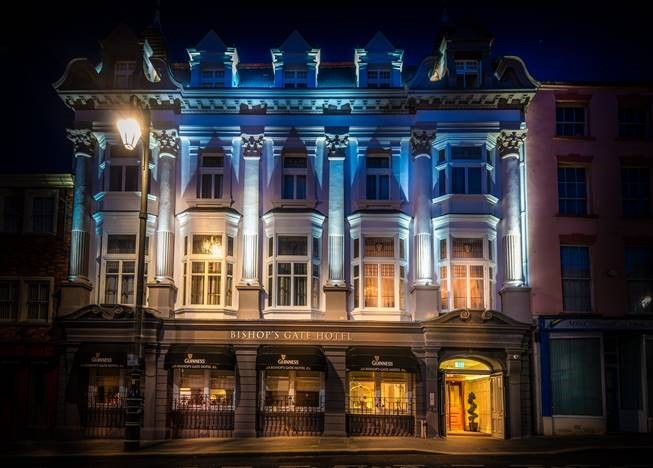 De sierlijke gevel van het Bishop's Gate Hotel, 's avonds verlicht, is voorzien van hoge ramen, decoratieve zuilen en zachtblauwe verlichting. Op de begane grond hangen Guinness-borden en warm verlichte entrees, die contrasteren met de donkere straat buiten. | Echt Ierland Vakanties