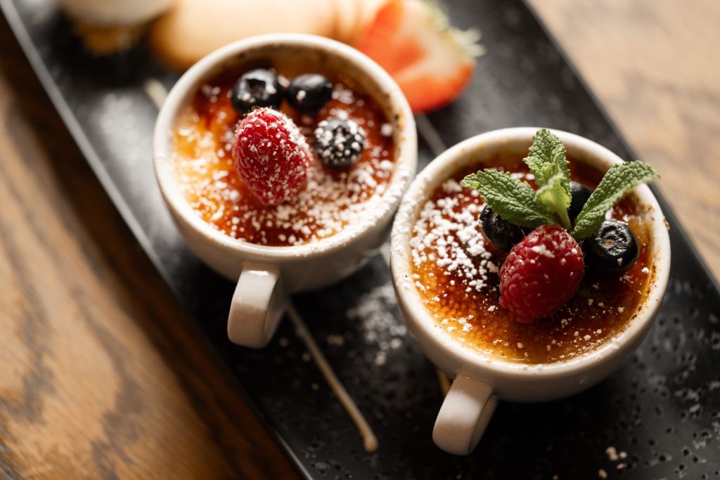 Twee kleine kopjes crème brûlée met poedersuiker, een framboos en bosbessen. Eén met een takje verse munt. Beide worden geserveerd op een zwart rechthoekig bord met een schijfje aardbei en een koekje op de wazige achtergrond. | Echt Ierland Vakanties
