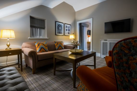 Een gezellige woonkamer met een bruine bank, kussens met oranje dessin en een bijpassende fauteuil. Een houten salontafel met een vaas en een mand. Zachte verlichting van lampen, ingelijste kunst aan de muur en een tv bij de deur zorgen voor extra comfort. De vloer is bekleed met een geruit tapijt. | Echt Ierland Vakanties