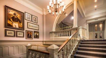 Elegante trap met witte balustrades en een gestreepte loper, die leidt naar een overloop met ingelijste portretten en kunstwerken aan beige muren. Een klassieke kroonluchter hangt erboven en rechtsboven de trap zijn glazen deuren zichtbaar. | Echt Ierland Vakanties