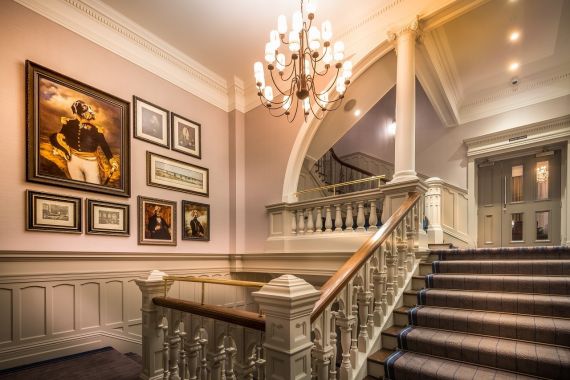 Elegante trap met witte balustrades en een gestreepte loper, die leidt naar een overloop met ingelijste portretten en kunstwerken aan beige muren. Een klassieke kroonluchter hangt erboven en rechtsboven de trap zijn glazen deuren zichtbaar. | Echt Ierland Vakanties