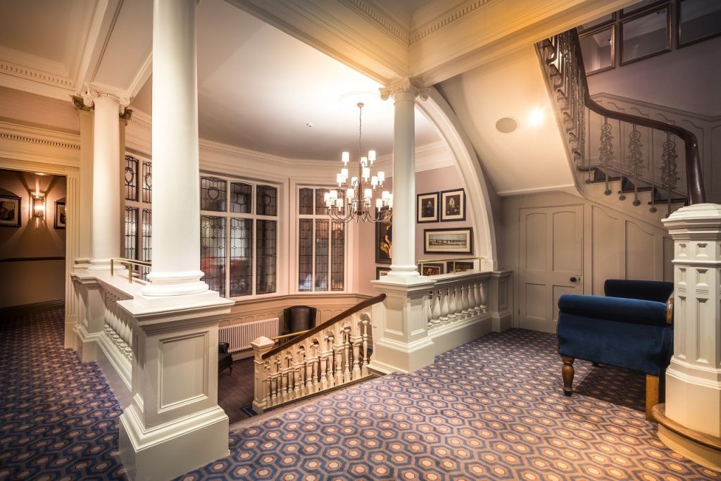 Elegant interieur met sierlijke witte zuilen, trapleuningen en sierlijsten. Een tapijt met een patroon bedekt de vloer en een blauwe fauteuil staat bij de trap. Muurkunst en een kroonluchter met meerdere lampen zorgen voor een klassieke touch. Glas-in-loodramen sieren de achterste muur. | Echt Ierland Vakanties