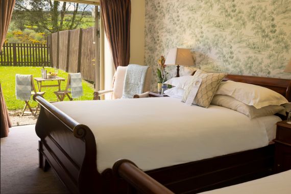 Een knusse slaapkamer met een houten slee-bed, opgemaakt met wit linnen en decoratieve kussens. Een lamp en stoel staan naast bloemenbehang. Glazen deuren geven toegang tot een zonnig terras met een tafel, stoelen, handdoeken en een groene tuin, omheind met hout. | Echt Ierland Vakanties
