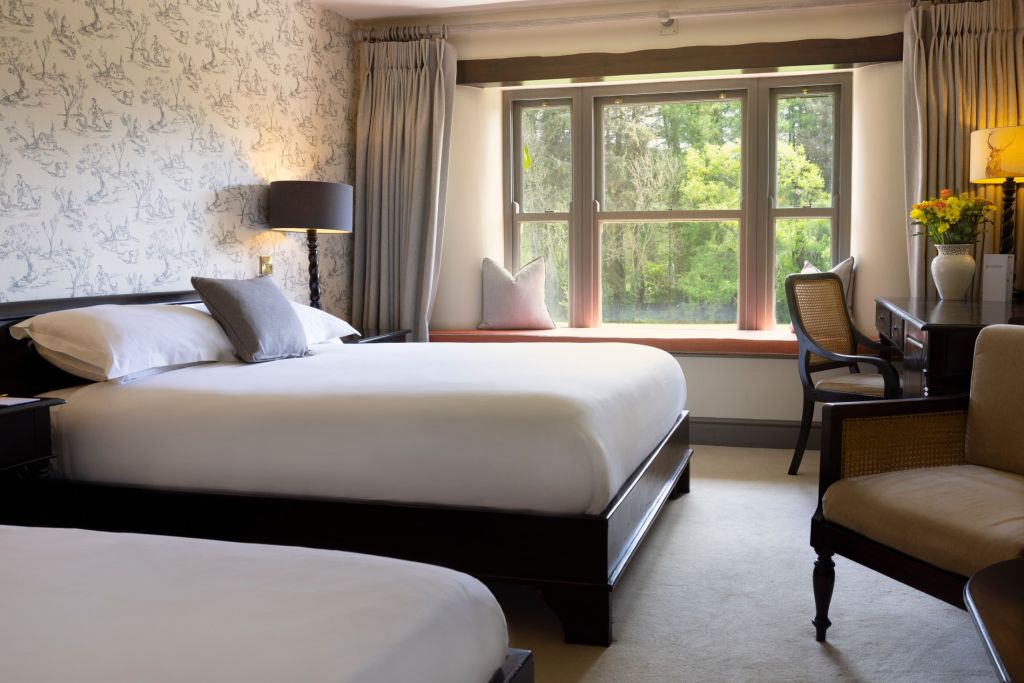 Lichte hotelkamer met twee tweepersoonsbedden, grijs en wit beddengoed, een vensterbank met kussens, een bureau met stoel, een lamp, behang met een subtiel patroon, een crèmekleurig tapijt en een vaas met bloemen op een bijzettafeltje. Buiten het raam zijn groene bomen te zien. | Echt Ierland Vakanties
