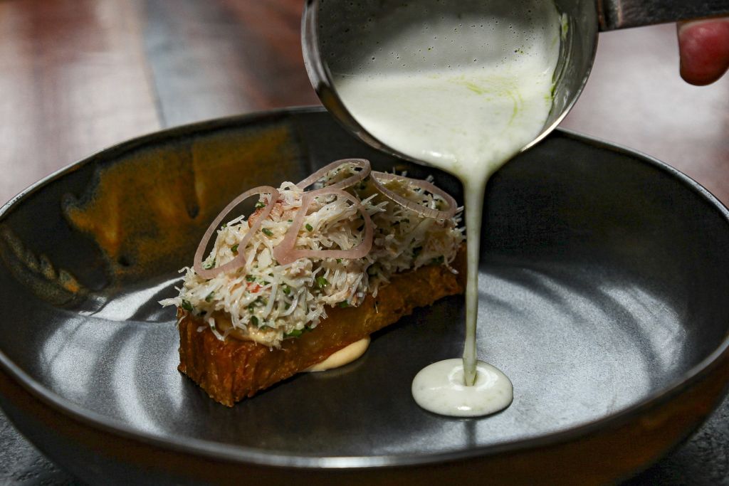 Een hand giet een lichtgroene, romige saus uit een metalen beker op een donker bord met daarop een rechthoekig stuk geroosterd brood, belegd met geraspte krab, dun gesneden sjalotjes en kruiden. Een klodder saus is gedeeltelijk verborgen onder de toast. | Echt Ierland Vakanties