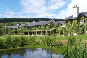 Een groot landhuis bedekt met groene klimop staat achter een vijver met riet. Het gebouw heeft witte schoorstenen, veel ramen en is omgeven door groen gras en bomen onder een gedeeltelijk bewolkte hemel. Beboste heuvels rijzen op de achtergrond. | Echt Ierland Vakanties