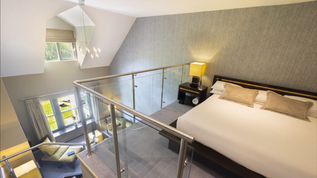 Een moderne loftslaapkamer met een groot bed, twee beige kussens, een nachtkastje met een lamp en een glazen balustrade die uitkijkt op een lichte woonkamer met ramen, gordijnen en een bank met kussens. Lichtgrijze muren en tapijt creëren een serene sfeer. | Echt Ierland Vakanties