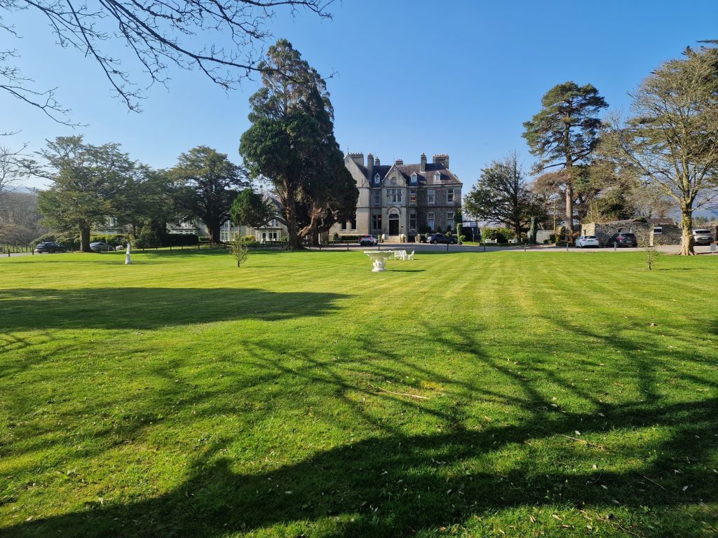 Een groot grasveld met nette strepen leidt naar een statig grijs stenen landhuis. Hoge bomen omringen het huis en werpen lange schaduwen. Een paar bankjes en sculpturen staan verspreid over het gazon. De lucht is helderblauw en er staan verschillende auto's bij het landhuis geparkeerd. | Echt Ierland Vakanties