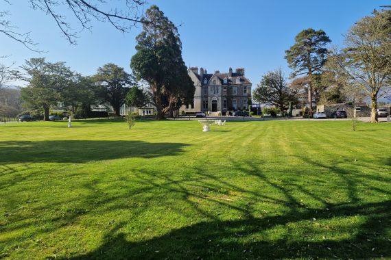 Een groot grasveld met nette strepen leidt naar een statig grijs stenen landhuis. Hoge bomen omringen het huis en werpen lange schaduwen. Een paar bankjes en sculpturen staan verspreid over het gazon. De lucht is helderblauw en er staan verschillende auto's bij het landhuis geparkeerd. | Echt Ierland Vakanties