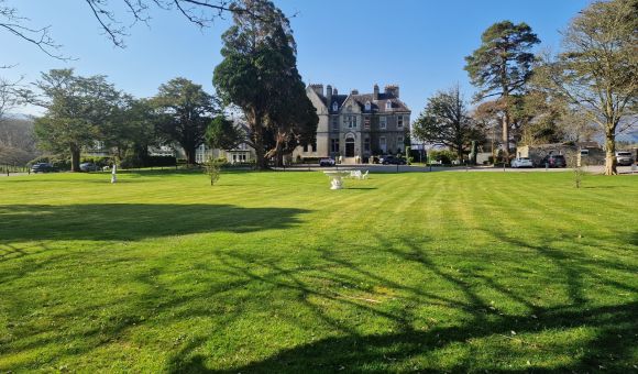 Een groot grasveld met nette strepen leidt naar een statig grijs stenen landhuis. Hoge bomen omringen het huis en werpen lange schaduwen. Een paar bankjes en sculpturen staan verspreid over het gazon. De lucht is helderblauw en er staan verschillende auto's bij het landhuis geparkeerd. | Echt Ierland Vakanties