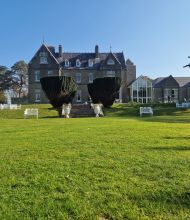 Een groot, grijs stenen landhuis met zadeldaken staat op een verzorgd groen gazon. Witte banken en stoelen staan verspreid over het gazon. Hoge, gesnoeide struiken flankeren een stenen trap die naar de ingang leidt. Bomen en een helderblauwe lucht vormen de achtergrond. | Echt Ierland Vakanties