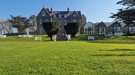 Een groot, grijs stenen landhuis met zadeldaken staat op een verzorgd groen gazon. Witte banken en stoelen staan verspreid over het gazon. Hoge, gesnoeide struiken flankeren een stenen trap die naar de ingang leidt. Bomen en een helderblauwe lucht vormen de achtergrond. | Echt Ierland Vakanties