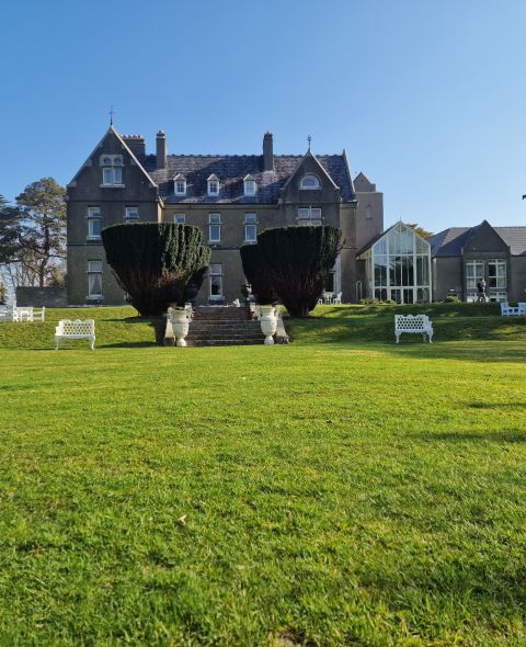Een groot, grijs stenen landhuis met zadeldaken staat op een verzorgd groen gazon. Witte banken en stoelen staan verspreid over het gazon. Hoge, gesnoeide struiken flankeren een stenen trap die naar de ingang leidt. Bomen en een helderblauwe lucht vormen de achtergrond. | Echt Ierland Vakanties
