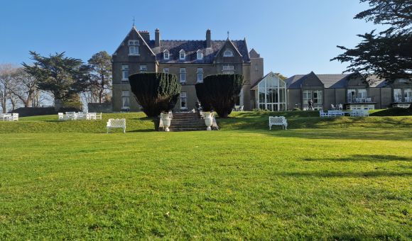 Een groot, grijs stenen landhuis met zadeldaken staat op een verzorgd groen gazon. Witte banken en stoelen staan verspreid over het gazon. Hoge, gesnoeide struiken flankeren een stenen trap die naar de ingang leidt. Bomen en een helderblauwe lucht vormen de achtergrond. | Echt Ierland Vakanties