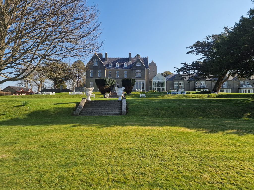 Op de achtergrond staat een groot, elegant landhuis, omringd door bomen. Een breed gazon leidt naar een stenen trap met witte beelden. De lucht is helderblauw en aan de rechterkant van het gebouw zijn moderne glazen uitbouwen zichtbaar. | Echt Ierland Vakanties