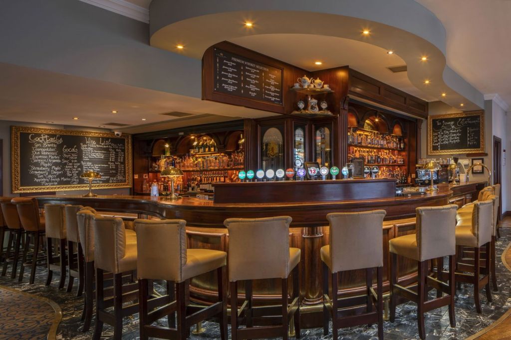 Een gepolijste houten bar met een gebogen toonbank wordt omringd door barkrukken met beige bekleding. Achter de bar staan rijen glazen en flessen netjes opgesteld. Aan de muur hangen menuborden met witte krijtletters. Warme verlichting zorgt voor een gezellige sfeer. | Echt Ierland Vakanties