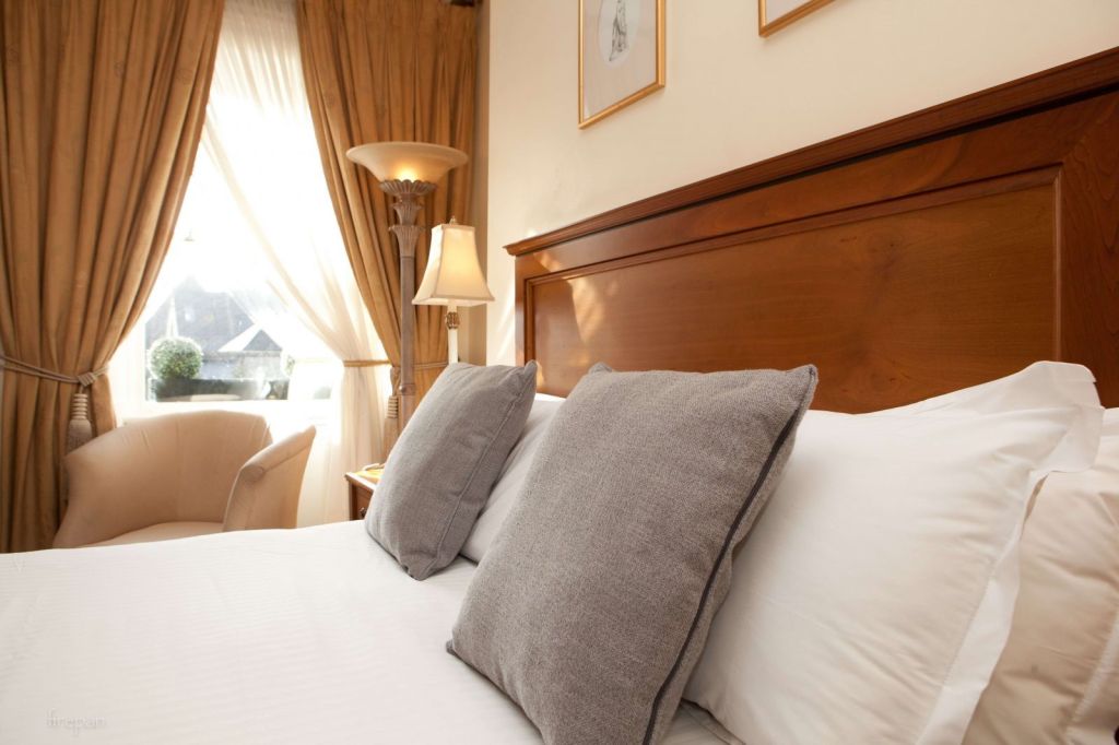 Een netjes opgemaakt bed met witte lakens en twee grote grijze kussens staat in een lichte kamer. Er is een houten hoofdeinde, beige gordijnen omlijsten een zonnig raam, een crèmekleurige fauteuil en warm licht van een klassieke tafellamp naast het bed. | Echt Ierland Vakanties