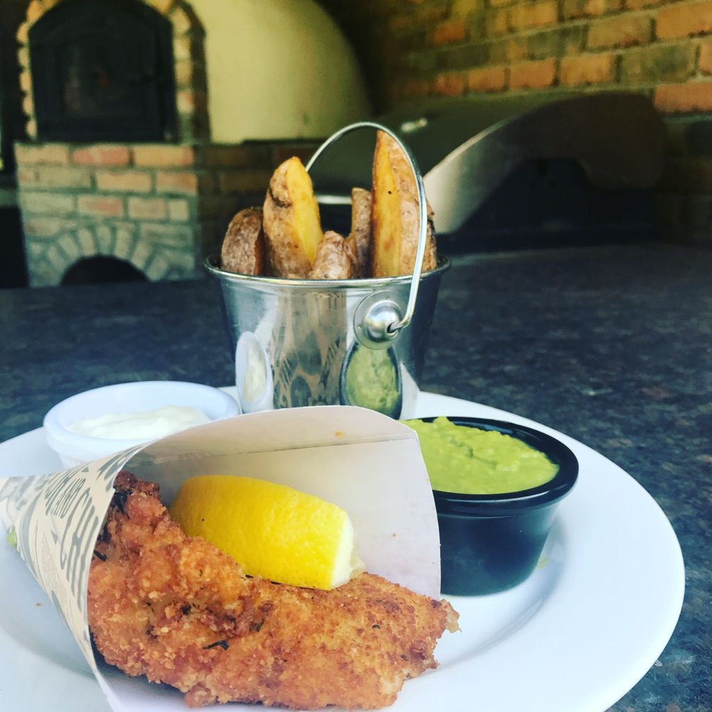 Een bord met gepaneerde vis en een citroenpartje in papier gewikkeld, geserveerd met een portie groene erwtenpuree en witte saus. Daarachter staat een kleine metalen emmer met dikke aardappelpartjes. Een steenoven en barbecue op de wazige achtergrond. | Echt Ierland Vakanties