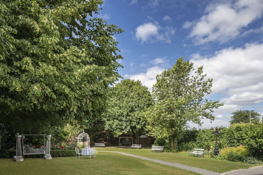 Een weelderige tuin met groene gazons, grote bomen en een kronkelend pad. Links staat een wit prieel met een tafel en stoelen klaar voor een kopje thee. Er staan verschillende witte bankjes langs de rand en de lucht is helder met hier en daar een wolkje. | Echt Ierland Vakanties