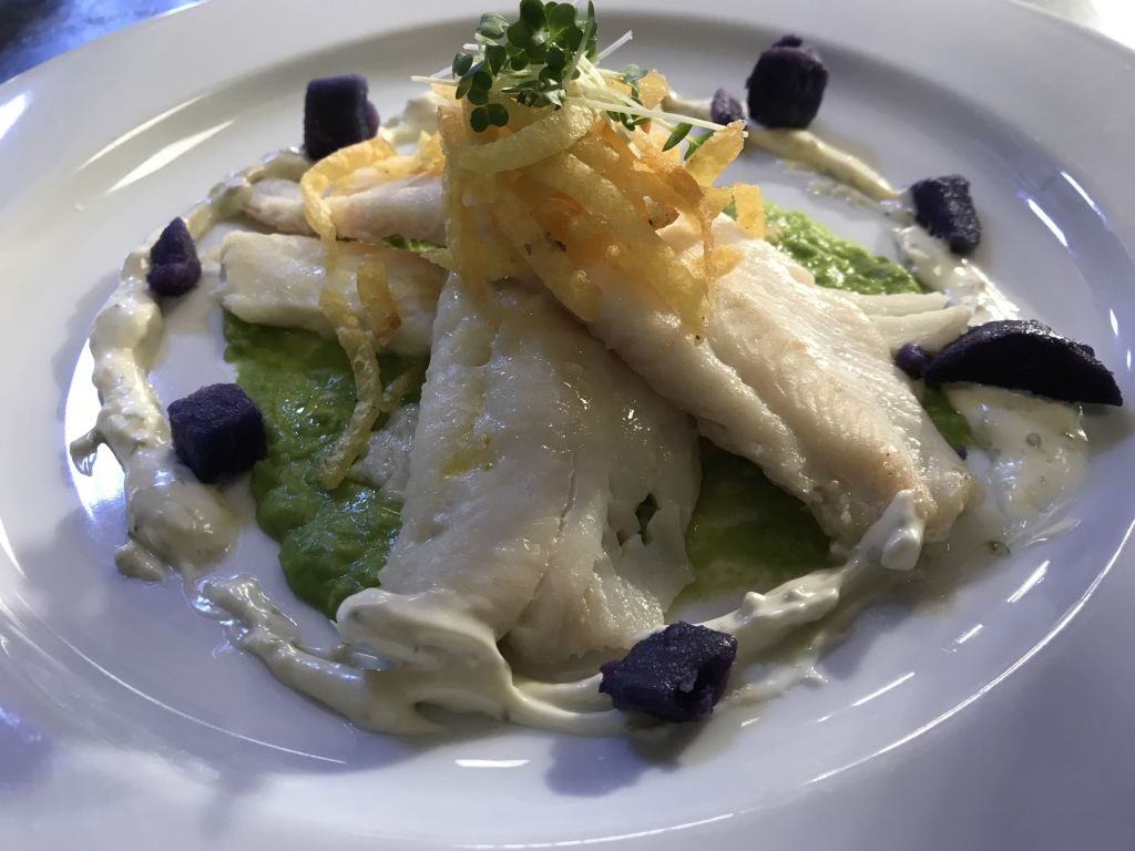 Een bord met witte visfilets op erwtenpuree, gegarneerd met knapperige, gefrituurde aardappelreepjes, microgroenten, een lepel romige witte saus en stukjes paarse aardappel, alles kunstig gepresenteerd op een wit bord. | Echt Ierland Vakanties