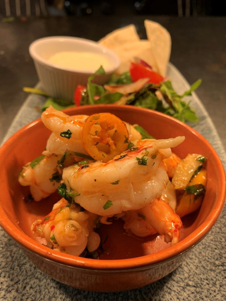 Een close-up van gekookte garnalen, gegarneerd met kruiden en een plakje chilipeper in een bruine kom. Op de achtergrond staat een klein schaaltje romige saus, verse salade met groenten en tomaat, en gevouwen tortillachips op een rechthoekig bord. | Echt Ierland Vakanties