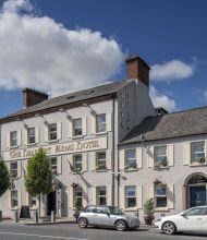 Een wit hotel en pub van drie verdiepingen met het uithangbord van The Charlemont Arms Hotel, hangende bloemenmanden en geparkeerde auto's. De lucht is blauw van de wolken en bomen staan langs de stoep voor het gebouw. | Echt Ierland Vakanties