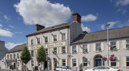 Een wit hotel en pub van drie verdiepingen met het uithangbord van The Charlemont Arms Hotel, hangende bloemenmanden en geparkeerde auto's. De lucht is blauw van de wolken en bomen staan langs de stoep voor het gebouw. | Echt Ierland Vakanties