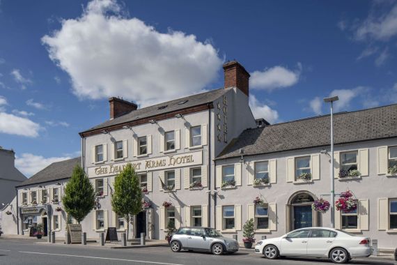 Een wit hotel en pub van drie verdiepingen met het uithangbord van The Charlemont Arms Hotel, hangende bloemenmanden en geparkeerde auto's. De lucht is blauw van de wolken en bomen staan langs de stoep voor het gebouw. | Echt Ierland Vakanties