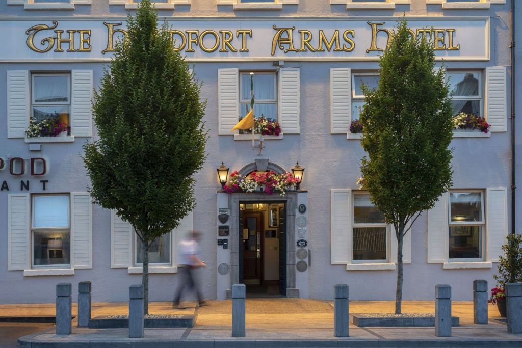 De gevel van The Headfort Arms Hotel is voorzien van crèmekleurige luiken, bloembakken en borden met gouden letters. Twee bomen omlijsten de ingang, waar een wazig persoon op de stoep langsloopt. Warm licht en potplanten accentueren de deuropening. | Echt Ierland Vakanties