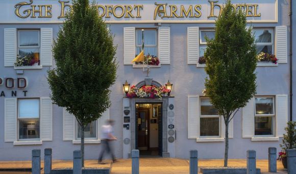 De gevel van The Headfort Arms Hotel is voorzien van crèmekleurige luiken, bloembakken en borden met gouden letters. Twee bomen omlijsten de ingang, waar een wazig persoon op de stoep langsloopt. Warm licht en potplanten accentueren de deuropening. | Echt Ierland Vakanties