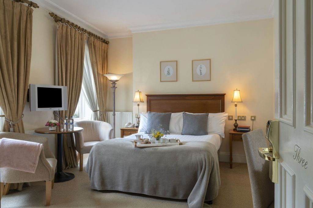 Een knusse hotelkamer met een tweepersoonsbed, beige muren, taupe gordijnen, een kleine ronde tafel met waterflesjes en glazen, twee stoelen, een flatscreen-tv, bedlampjes en ingelijste kunst boven het bed. Op het bed staat een dienblad met thee en bloemen. | Echt Ierland Vakanties