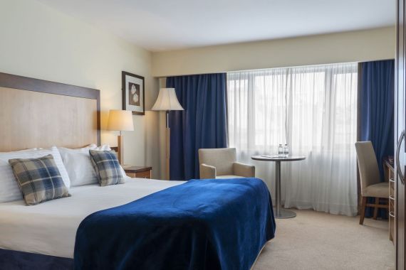 Een netjes ingerichte hotelkamer heeft een tweepersoonsbed met een blauwe deken en geruite kussens, een houten hoofdeinde, een nachtkastje met lamp, twee fauteuils, een ronde tafel met twee waterflessen, een staande lamp, een raam met blauwe gordijnen en een ingelijste kunst aan een crèmekleurige muur. | Echt Ierland Vakanties