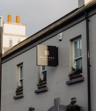 Een beige gebouw met bloembakken en een zwart bord met de tekst Hillyard House in gouden letters hangt boven de ingang. Het gebouw heeft een schuin dak en schoorstenen, met een lantaarnpaal en een heldere hemel op de achtergrond. | Echt Ierland Vakanties
