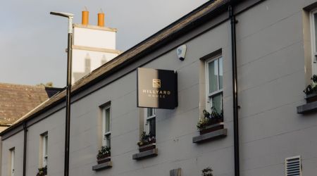 Een beige gebouw met bloembakken en een zwart bord met de tekst Hillyard House in gouden letters hangt boven de ingang. Het gebouw heeft een schuin dak en schoorstenen, met een lantaarnpaal en een heldere hemel op de achtergrond. | Echt Ierland Vakanties