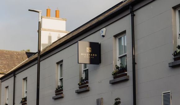 Een beige gebouw met bloembakken en een zwart bord met de tekst Hillyard House in gouden letters hangt boven de ingang. Het gebouw heeft een schuin dak en schoorstenen, met een lantaarnpaal en een heldere hemel op de achtergrond. | Echt Ierland Vakanties
