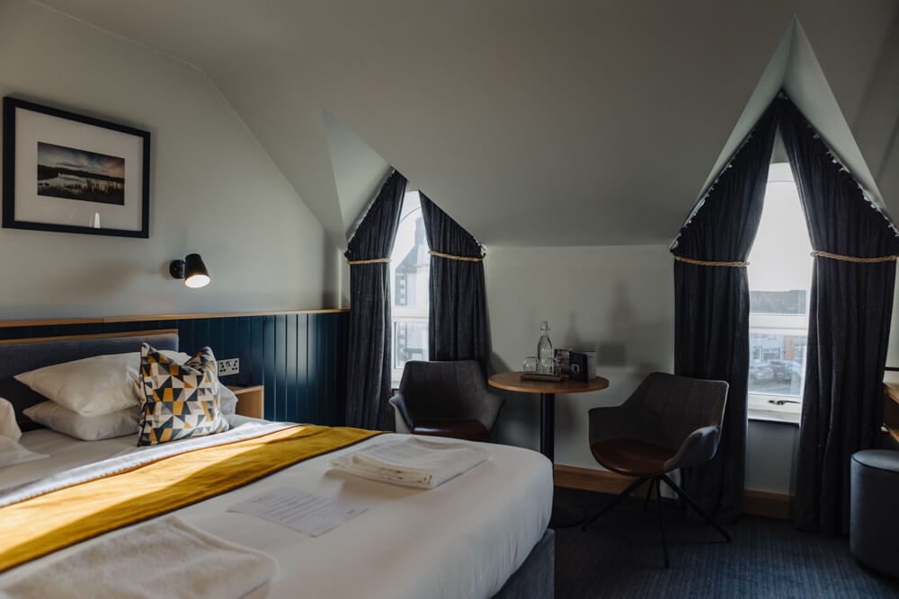 Een knusse hotelkamer met schuine plafonds, een tweepersoonsbed met wit linnen en een gele deken, twee geometrische kussens, een kleine ronde tafel met twee stoelen, een fles water en twee hoge ramen met donkere gordijnen. Een ingelijste foto hangt aan de muur. | Echt Ierland Vakanties