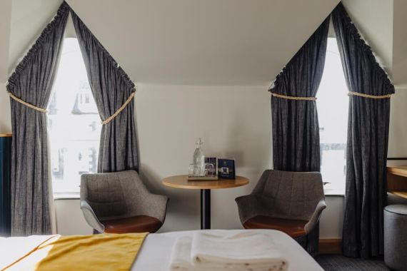 Een knusse hotelkamer met twee grijze fauteuils rond een kleine ronde tafel met flesjes water en glazen. Twee boogramen met donkere gordijnen verlichten de ruimte. Op de voorgrond is een bed met wit linnen en een gele loper zichtbaar. | Echt Ierland Vakanties