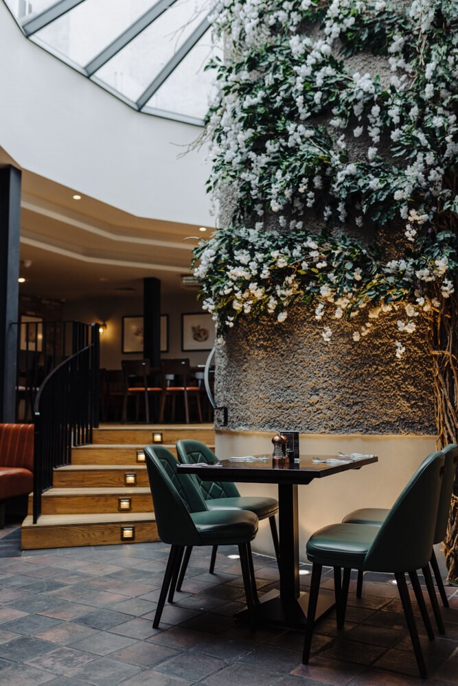 Een gezellig restaurantinterieur met groene stoelen rond een zwarte tafel, gedekt voor twee personen. Een muur is bedekt met weelderige witte bloemen en groen. Een houten trap met kleine lampjes leidt naar een eetgedeelte op de bovenverdieping. Een dakraam laat natuurlijk licht binnen. | Echt Ierland Vakanties