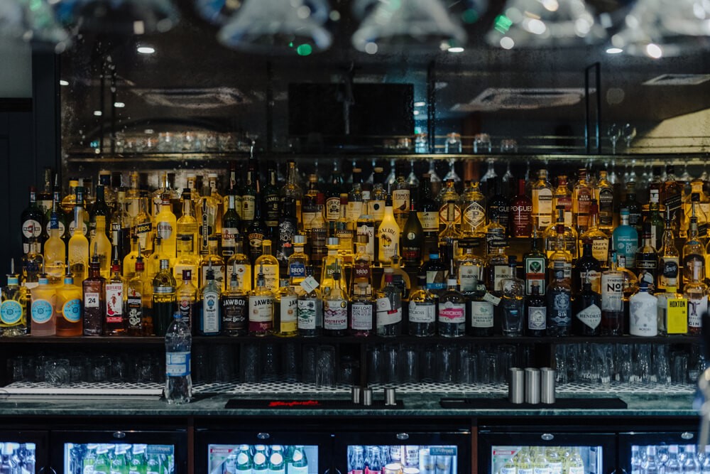 Een barplank vol met diverse flessen sterke drank, waaronder whisky, wodka, gin, rum en likeuren. De flessen staan dicht op elkaar en worden van achteren belicht. Eronder zijn glaswerk en gekoelde drankenvitrines zichtbaar. | Echt Ierland Vakanties