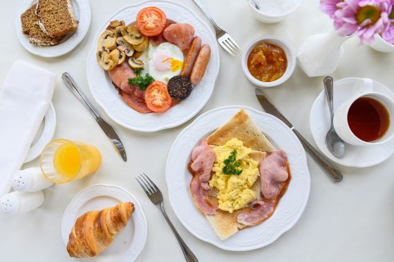 Een gedekte tafel voor het ontbijt met borden vol eieren, spek, worst, gegrilde tomaat, champignons, bloedworst, toast, een croissant, sinaasappelsap, thee, marmelade en bruin brood. Een roze bloem in een vaas en bestek liggen ook op het witte tafelkleed. | Echt Ierland Vakanties
