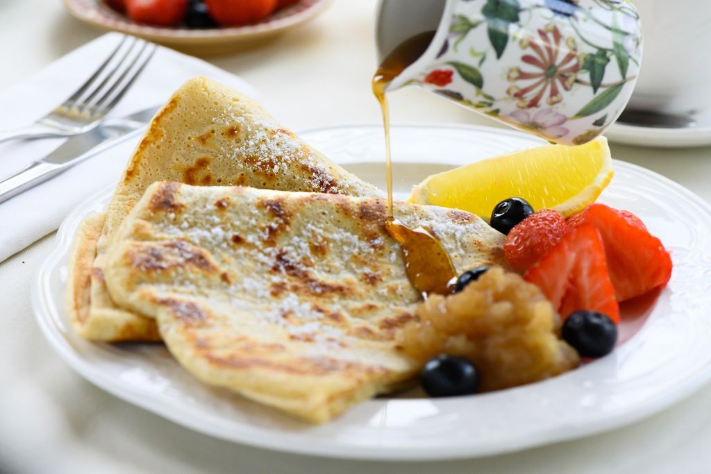 Een bord met gevouwen crêpes, bestrooid met poedersuiker, geserveerd met siroop, appelcompote, verse aardbeien, bosbessen en een schijfje sinaasappel. Op de achtergrond zijn een vork en mes te zien, samen met een siroopkan met bloemmotief. | Echt Ierland Vakanties